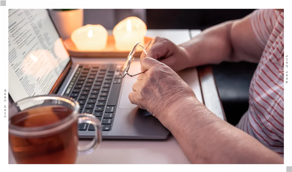 An older person's hands resting on a laptop keyboard in a cozy, candlelit setting with a cup of tea nearby — suggesting a calm, unhurried moment of taking care of important personal matters.